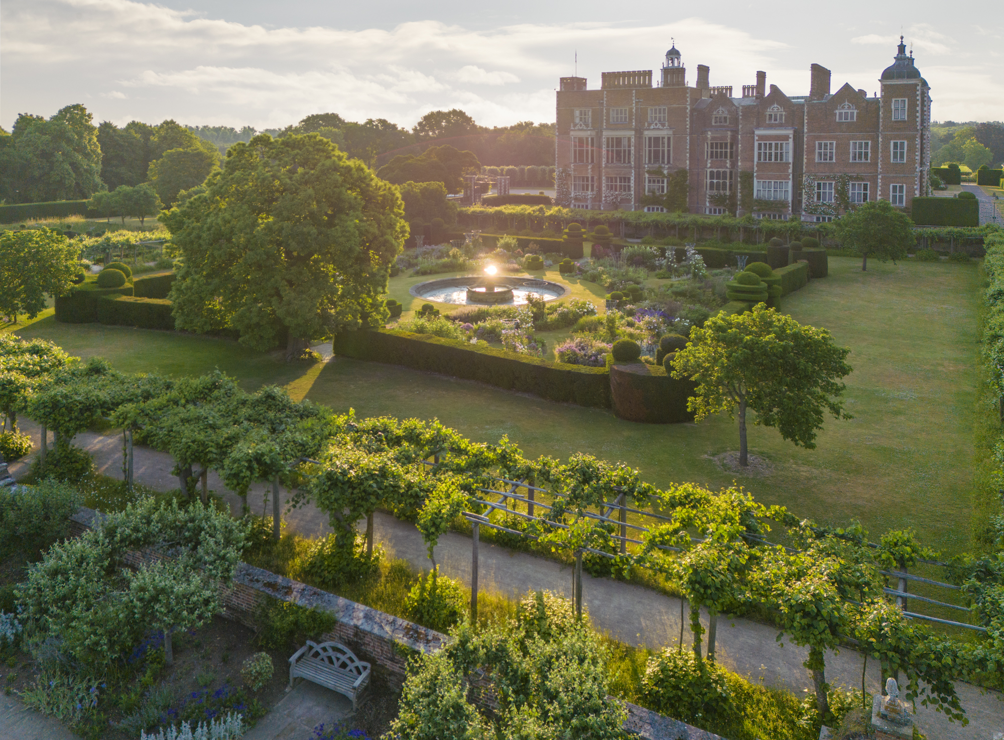 Aerial view of a historic mansion surrounded by manicured gardens. A fountain glows in sunlight, with lush trees and hedges under a serene sky.